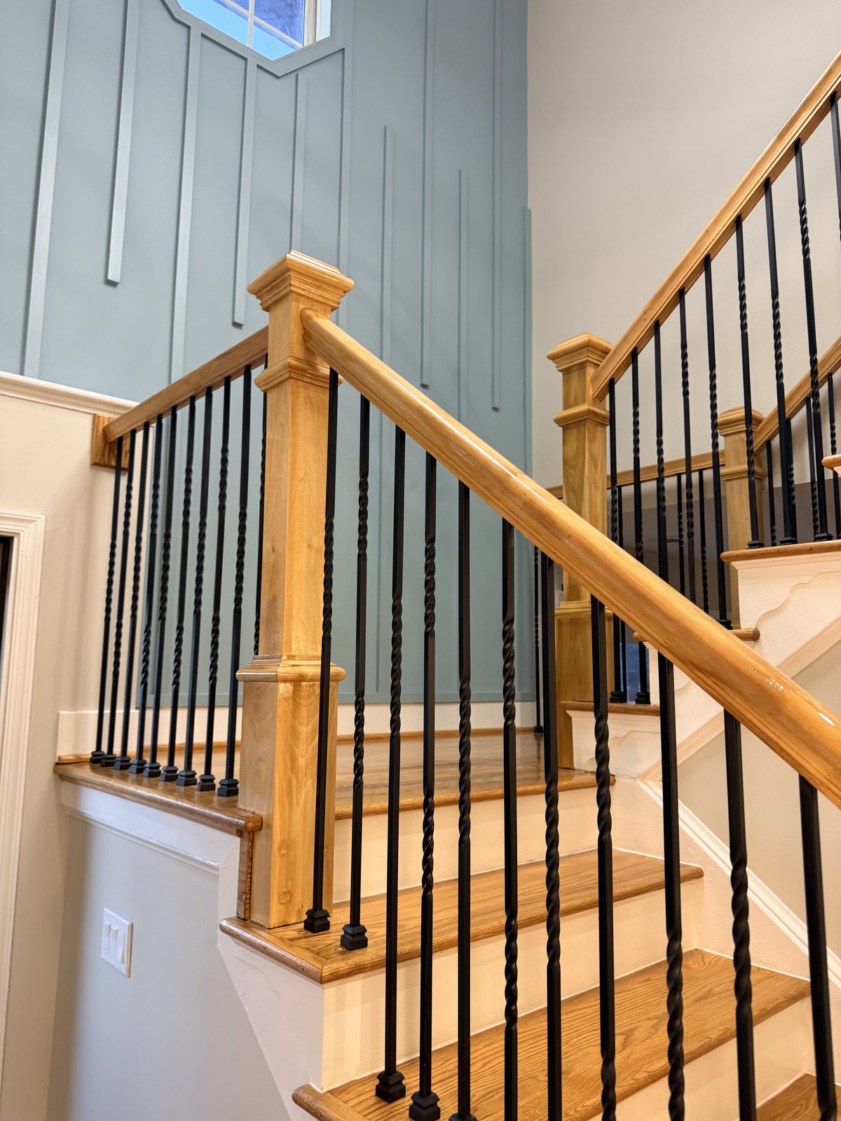 Refinished oak handrail and newel post with black iron balusters beside a blue board-and-batten wall